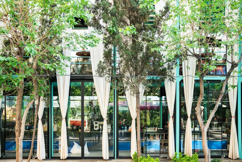 کافه‌حیات هزار افسانْ: نمای بیرونی مدرن کافه با قاب‌های فیروزه‌ای، شیشه‌های وسیع، پرده‌های سفید و درختان سرسبز جلوی ساختمان.