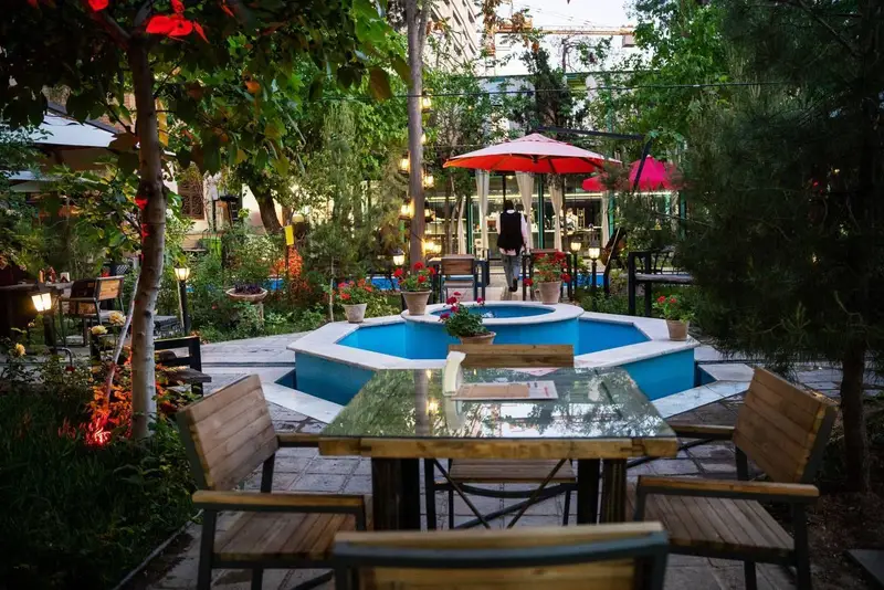 تصویری زیبا از فضای باز و باغ سنتی کافه‌حیات هزار افسانْ با حوض آبی مرکزی، نورپردازی دلنشین و میز و صندلی‌های چوبی.