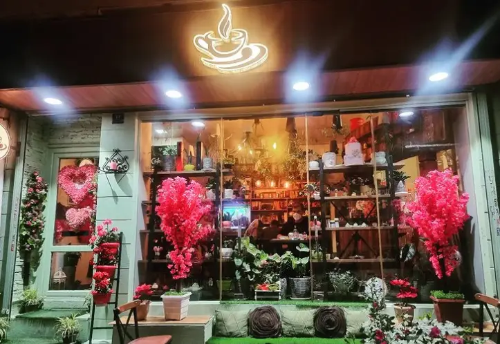 نمای بیرونی کافه دونه نارمک در شب با نورپردازی گرم و جذاب، تابلوی فنجان قهوه درخشان، ورودی تزئین شده با قلب‌های صورتی و پنجره‌ای بزرگ از فضای داخلی سرسبز و مملو از گل و گیاه زیبا