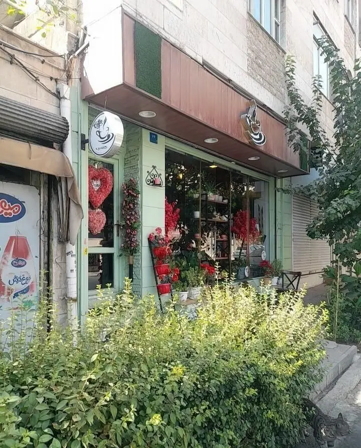 نمای بیرونی کافه دونه نارمک با نمای سبز و دکوراسیون گل و گیاه، ویترین پر از گل‌های قرمز، تابلوی جذاب کافه و بوته‌های سبز جلوی آن.