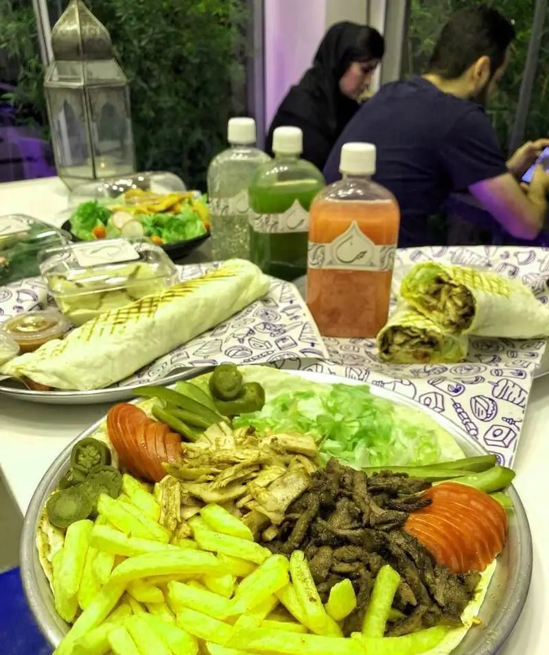 میز مملو از غذاهای فست فود حب: شاورما گوشت و مرغ، سیب زمینی، سالاد، ساندویچ و نوشیدنی در فضای رستوران.