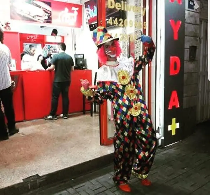 یک دلقک با لباس شاد و رنگارنگ و کلاه بانمک، جلوی ورودی فست فود پاندا. در داخل، مشتریان پشت کانتر قرمز سفارش می‌دهند.