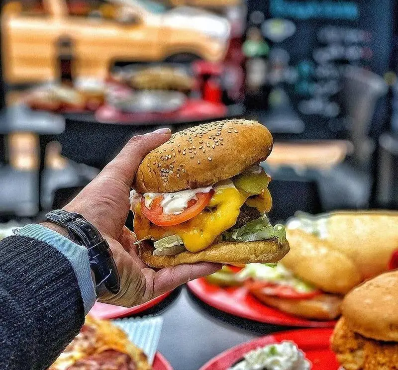 یک برگر ویژه و آبدار پنیر از فست فود پاندا در دست، با نان کنجدی، کاهو، گوجه، خیارشور و سس سفید، ظاهری جذاب و دلپذیر.