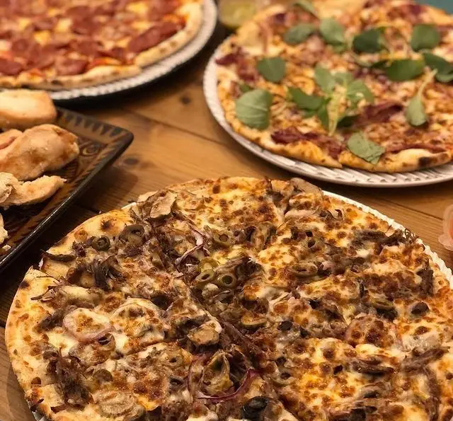 تصویری جذاب از سه پیتزای خوشمزه و داغ پاستا چنگال شامل پیتزا پپرونی، پیتزا قارچ و زیتون روی میز چوبی رستوران.
