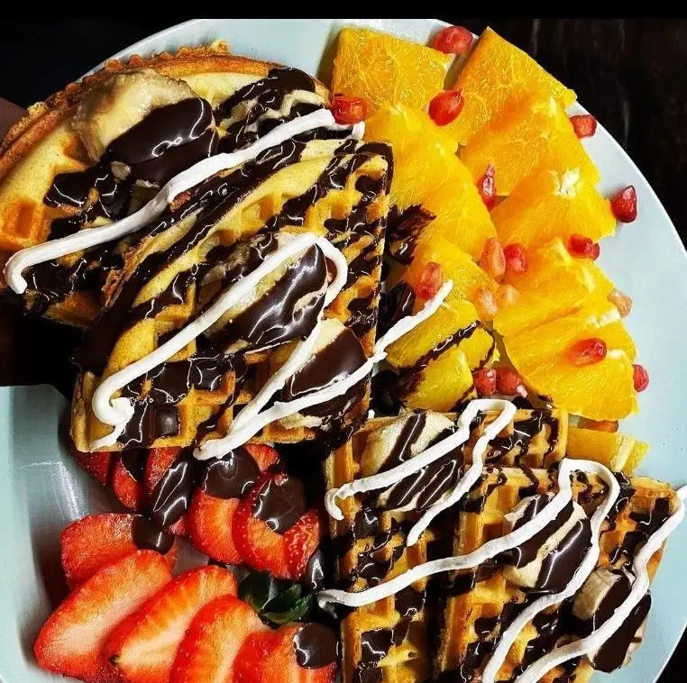 تصویر وافل‌های کافه اوتن با روکش سس شکلات و سس سفید، تزئین شده با توت فرنگی، پرتقال، موز و انار. یک دسر یا صبحانه لذیذ.