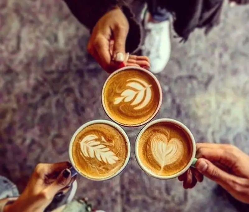 سه فنجان قهوه لاته آرت خلاقانه با طرح‌های برگ و قلب در دستان دوستان، انتخابی عالی برای دورهمی در کافه اوتن.