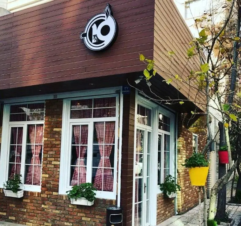 نمای بیرونی کافه اوتن در تهران، با دکوراسیون آجری و چوبی، پنجره‌های سفید با پرده‌های شطرنجی قرمز و گلدان‌های زیبا، فضایی دلنشین و دنج.