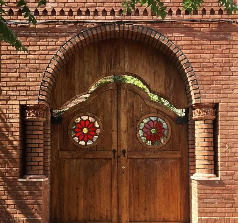 نمای ورودی عمارت نیلی با دروازه چوبی زیبا، طاق هلالی آجری، شیشه‌های رنگی گل‌دار و ستون‌های تزئینی سنتی.