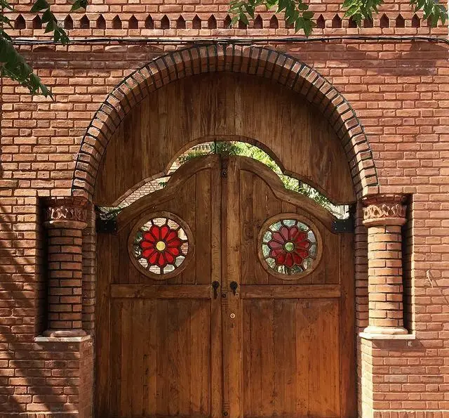 ورودی چوبی زیبای عمارت نیلی با طرح شیشه رنگی گل سرخ در دیوار آجری، نشانگر معماری سنتی و دلنشین ایرانی.