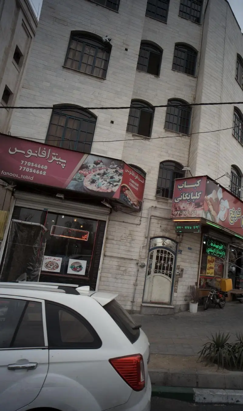 تصویری از نمای خارجی فست فود فانوس توحید، رستورانی در شهر، با تابلوهای پیتزا و ساندویچ، در کنار یک کباب فروشی در خیابان.