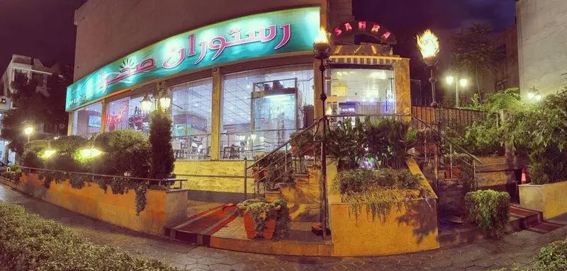 نمای شبانه و ورودی جذاب باغچه رستوران سنتی صحرا، با تابلو روشن، مشعل‌های فروزان، نورپردازی گرم و فضای سبز انبوه.