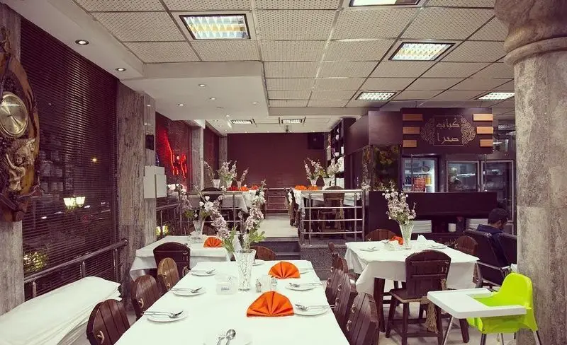 تصویری از داخل باغچه رستوران سنتی صحرا با میزهای چیده شده، گل و تزئینات، آماده پذیرایی از مهمانان برای تجربه غذای ایرانی اصیل.