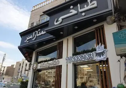 نمای بیرونی ساختمان طباخی ممتاز امیر، شامل تابلوهای فارسی و انگلیسی، شماره تلفن 77888283 و نماد دیگ غذا با شعله‌های آتش.