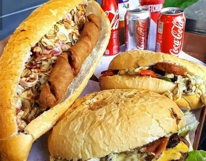 نمای نزدیک از انواع ساندویچ و برگر در یک فست فود: هات داگ غول‌پیکر با قارچ، دو برگر گوشت با پنیر و بیکن، و کوکاکولا