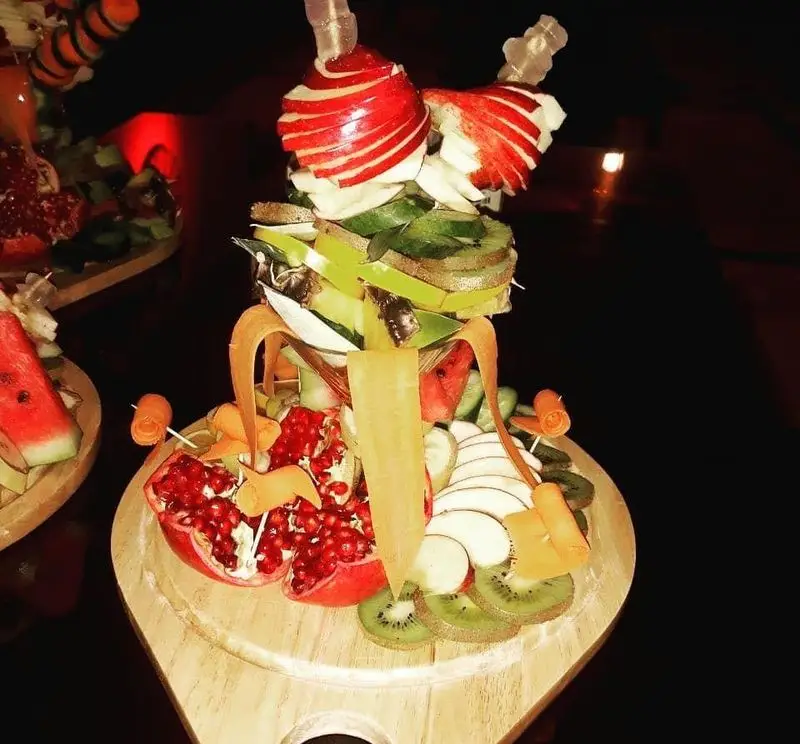 میوه آرایی باشکوه و خلاقانه در رستوران مروارید، شامل سیب حکاکی شده، کیوی، انار و هندوانه تازه روی سینی چوبی، ایده‌آل برای پذیرایی مهمانان.