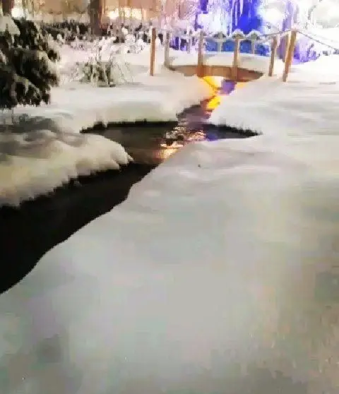 منظره‌ای سحرانگیز از رستوران مروارید در شبی برفی: رودخانه‌ای آرام، پل چوبی مزین، نورهای گرم منعکس در آب و درختان پوشیده از برف.