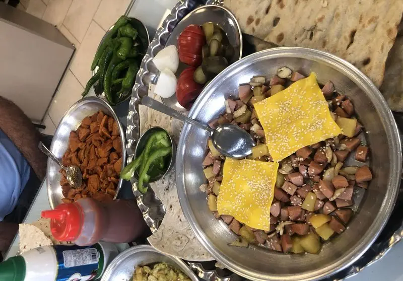 میز پر از غذاهای متنوع و خوشمزه کیان لقمه؛ بشقاب بزرگ سوسیس، سیب زمینی و پنیر به همراه نان و دورچین‌های تازه.