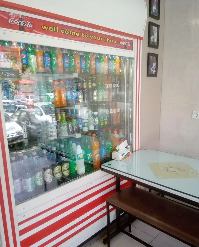 فروشگاه کیان لقمه با یخچال بزرگ و پر از نوشیدنی‌های متنوع شامل انواع نوشابه، آبمیوه و ماءالشعیر، و فضای دلپذیر با میز و نیمکت برای مشتریان.