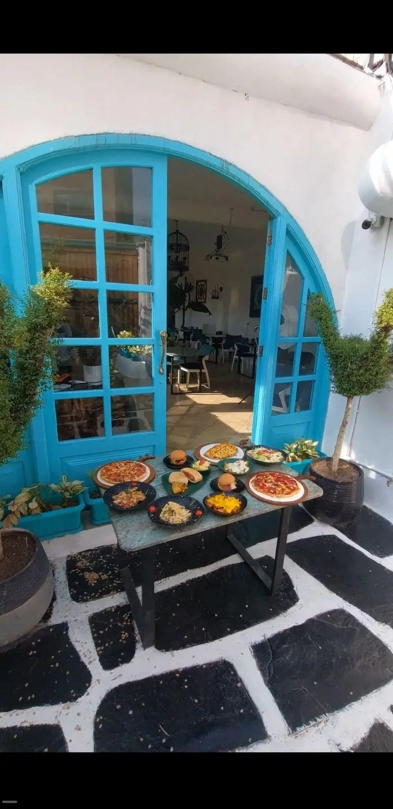 کافه رستوران حیات نو با در آبی روشن، میزی پر از پیتزا، برگر، پاستا و سالاد در فضای باز با کفپوش سیاه و سفید و گلدان.