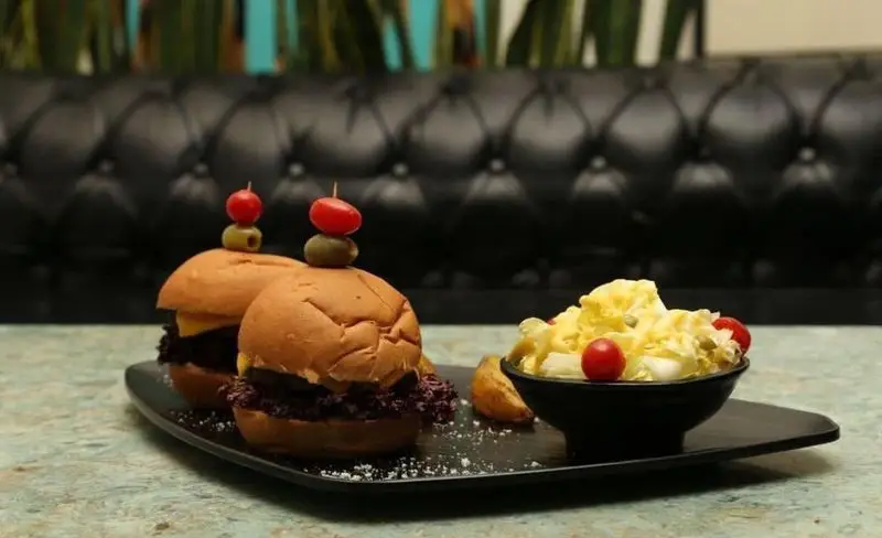 دو همبرگر کوچک با سس و پنیر به همراه سالاد و سیب‌زمینی در سینی مشکی کافه رستوران حیات نو. تجربه غذایی دلپذیر از منو.