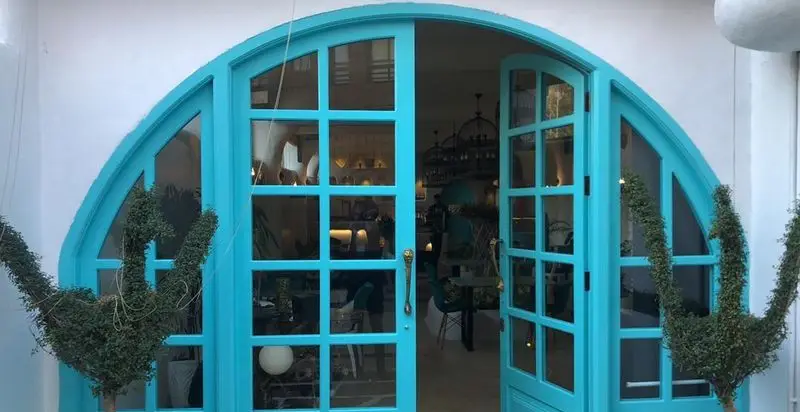 درب ورودی آبی فیروزه ای و طاق دار کافه رستوران حیات نو، با پنجره‌های مشبک و گیاهان زینتی خاص، نمایی دنج و دعوت کننده از فضای داخلی.