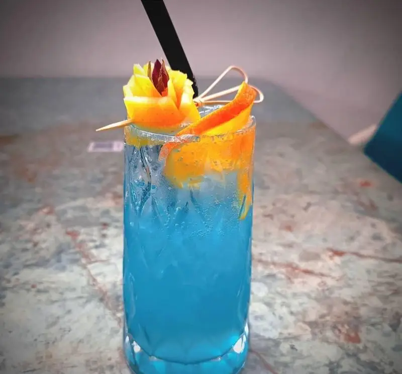 کوکتل آبی جذاب و خنک کافه رستوران حیات نو، تزئین شده با پوست پرتقال و نی مشکی، آماده لذت بردن در فضایی دلنشین.