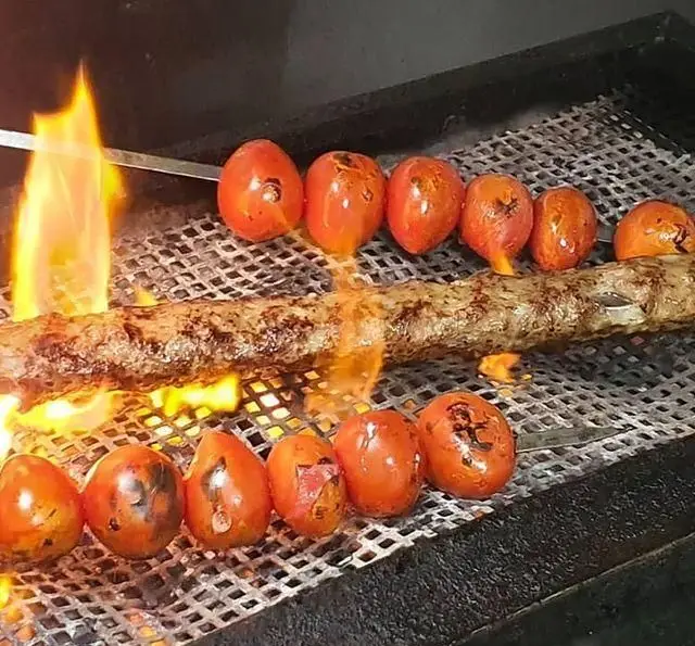 کباب کوبیده لذیذ و گوجه‌های کبابی آبدار در حال پخت روی گریل ذغالی با شعله‌های آتش در رستوران زیتون، تجربه‌ای فراموش نشدنی.