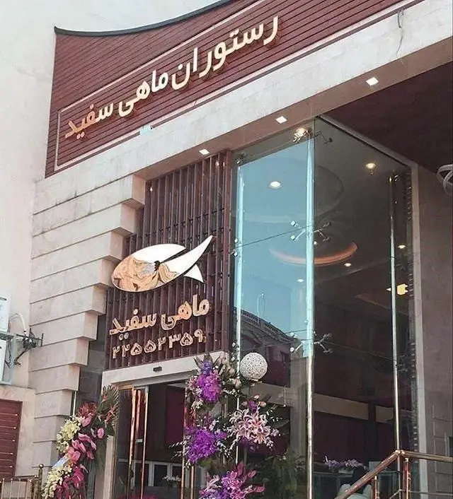 نمای بیرونی و ورودی اصلی رستوران ماهی سفید، تابلوی بزرگ با نام "رستوران ماهی سفید"، لوگوی ماهی طلایی و تزئینات گل‌های رنگی. محیطی دلنشین برای سرو غذای دریایی.