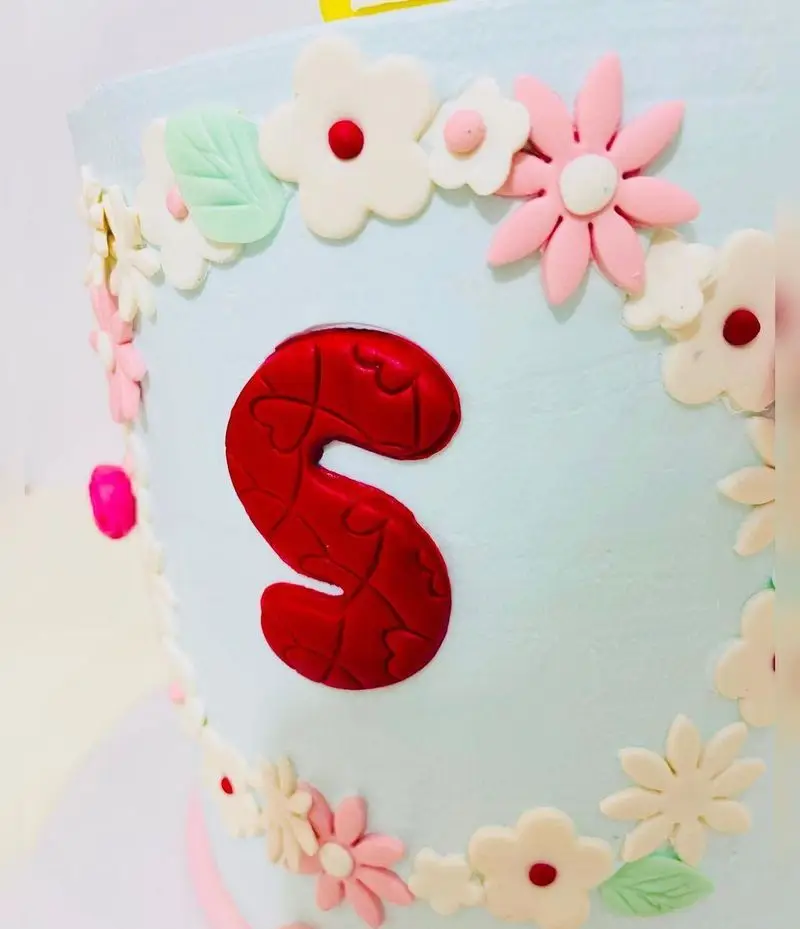 کیک فوندانت آبی روشن و زیبای قنادی نارملینو، تزیین شده با گل‌های بهاری صورتی و سفید، برگ‌های سبز و حرف S برجسته قرمز با طرح قلب.