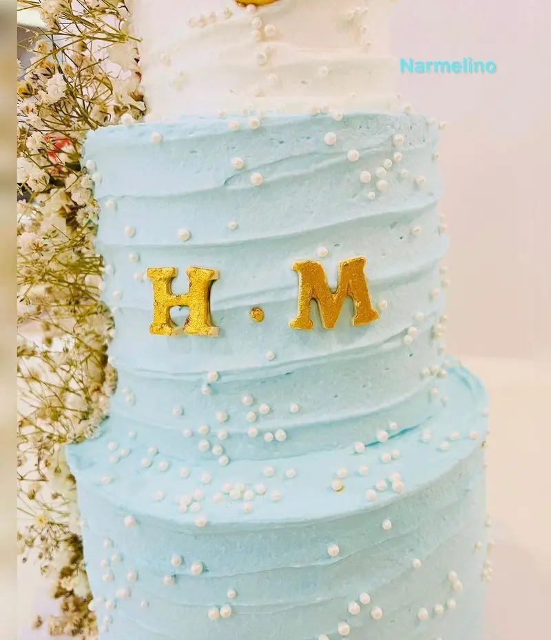 کیک تولد سه طبقه آبی و سفید شیک و سفارشی از قنادی نارملینو با حروف طلایی H.M، مناسب جشن ها و مراسم خاص.
