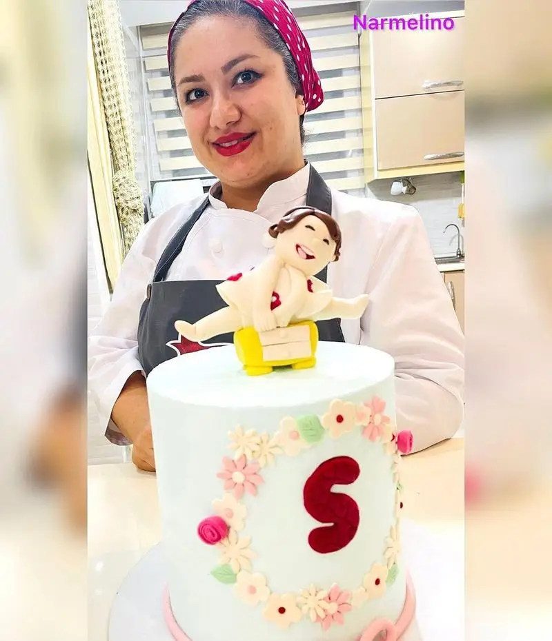 کیک تولد سفارشی قنادی نارملینو، با تزئین فوندانت گل، حرف S و فیگور ورزشی شاد. زن قناد با افتخار در کنار کارش.