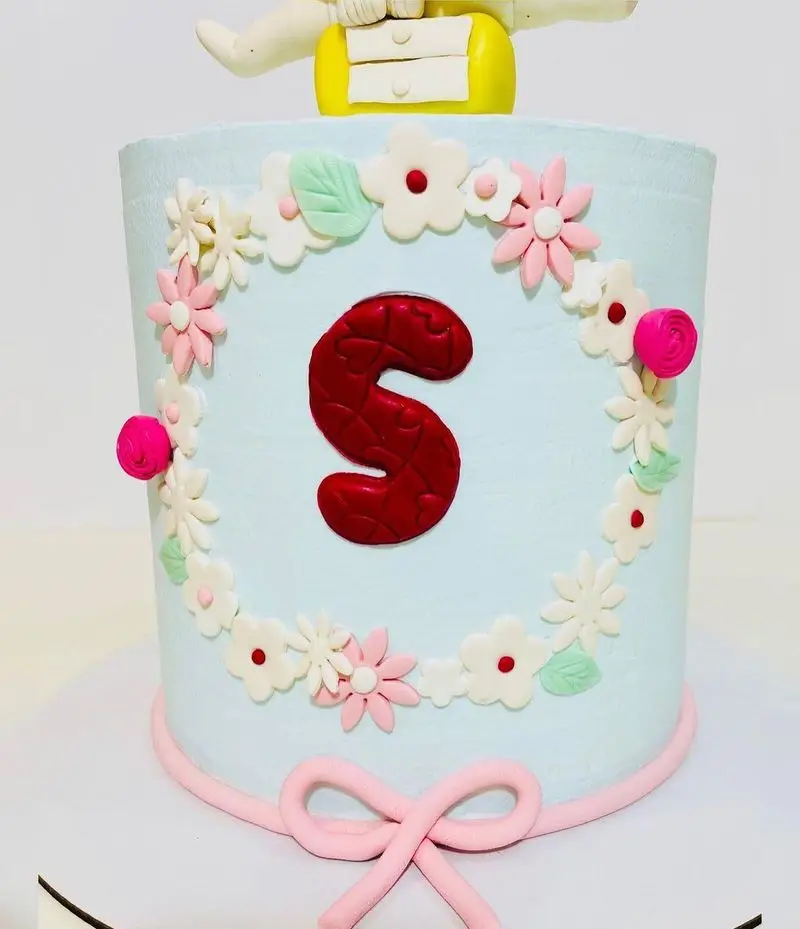 کیک تولد فوندانت آبی رنگ قنادی نارملینو با تزیین گل‌های صورتی و سفید، حرف S قرمز و پاپیون صورتی. مناسب جشن‌ها و سفارشات خاص.