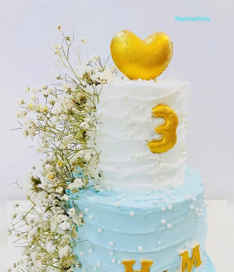 کیک جشن تولد سه سالگی نارملینو، دو طبقه با روکش سفید و آبی، تزیینات قلب و عدد طلایی و گل‌های سفید تازه برای خاطرات شیرین.