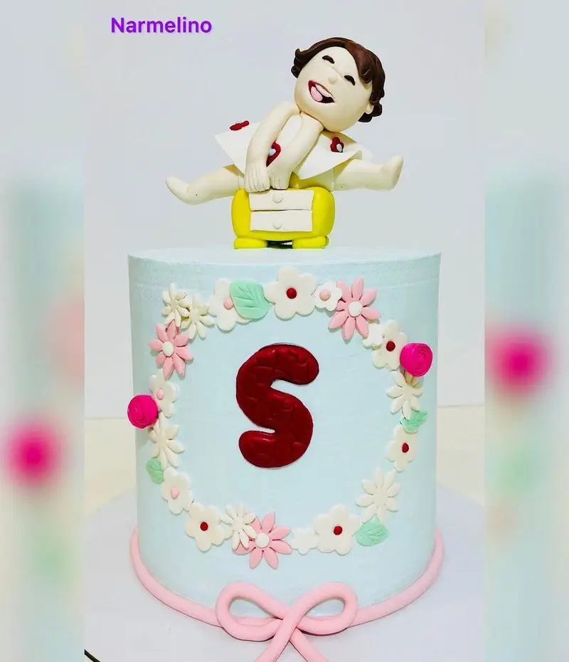 کیک تولد دخترانه آبی رنگ قنادی نارملینو با تاپر کودک خندان، حرف S قرمز برجسته و تزیینات زیبای گل و روبان صورتی.