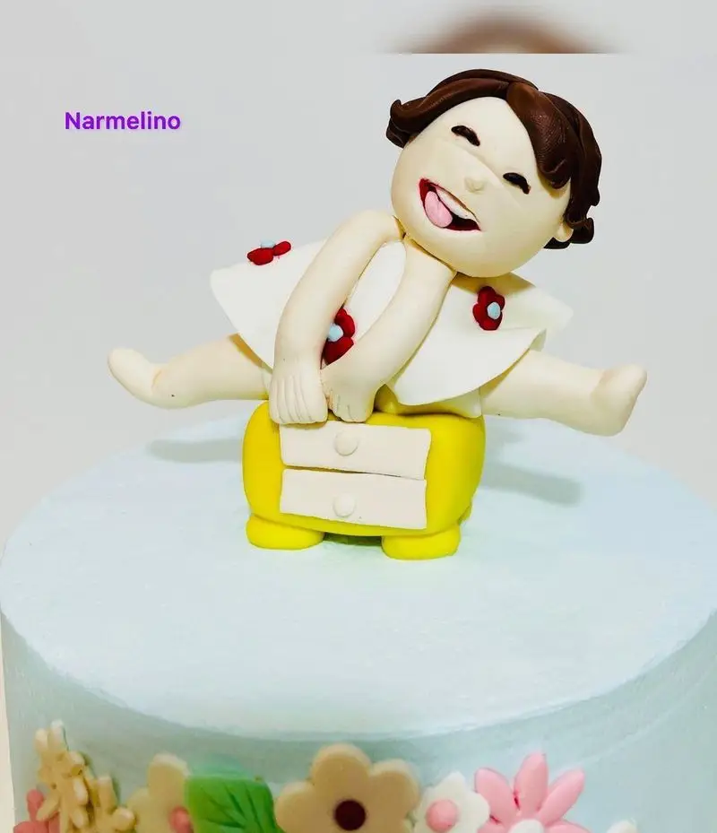 تاپر کیک فوندانت دست‌ساز و زیبای کودک خندان و شاد درازکش روی دراور زرد، ایده‌آل برای کیک تولد از قنادی نارملینو.