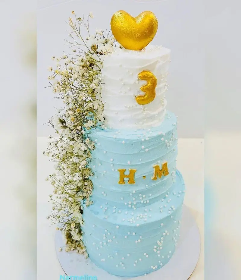 کیک تولد دو طبقه آبی و سفید قنادی نارملینو با تاپر قلب طلایی، عدد 3، حروف H.M، مرواریدهای خوراکی و گل‌های عروس.