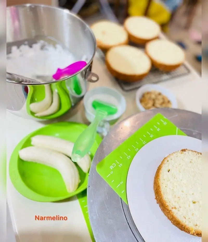 تصویری از مراحل آماده سازی کیک موز از قنادی نارملینو با مواد اولیه تازه شامل موز، خامه، و لایه های کیک اسفنجی روی میز کار.