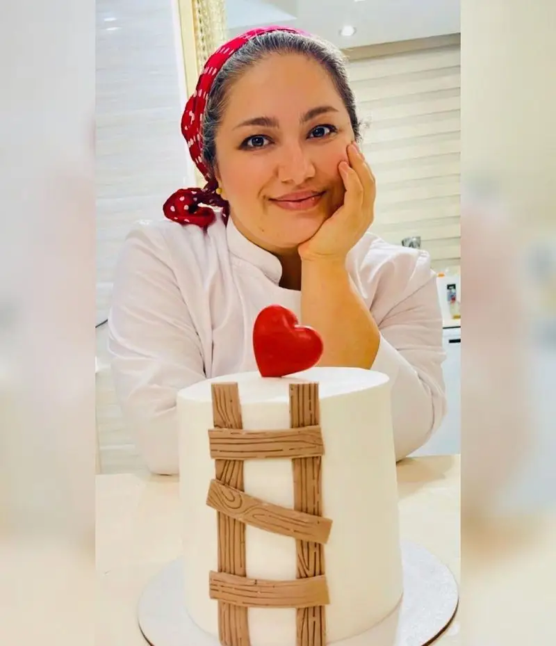 تصویر بانوی شیرینی‌پز قنادی نارملینو کنار کیک فانتزی با دیزاین منحصر به فرد قلب و چوب، طعم و زیبایی در یک قاب.
