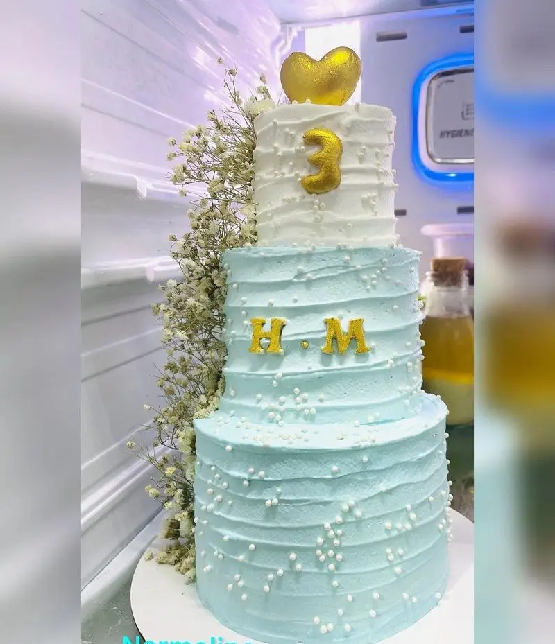 کیک تولد سه طبقه آبی و سفید از قنادی نارملینو. دارای تزیینات قلب طلایی، عدد 3، حروف H.M و گلهای ژیپسوفیلا.
