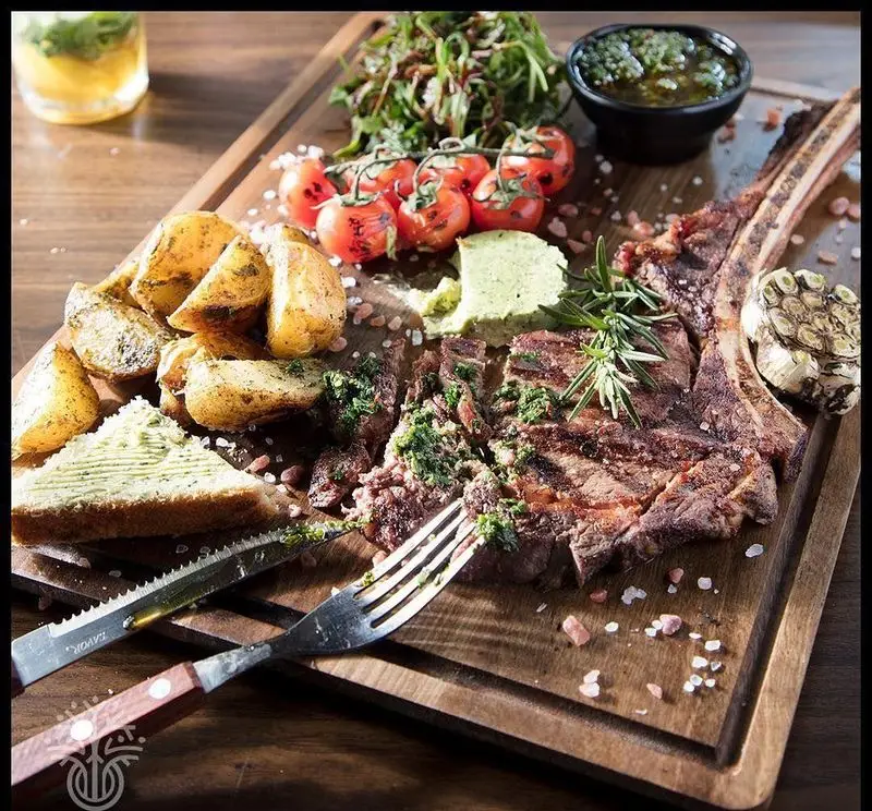 استیک تاماهاک گریل شده با سیب زمینی تنوری، گوجه فرنگی گیلاسی و سس چیمچوری در کافه رستوران لوکال ولنجک، یک غذای اصلی لذیذ.