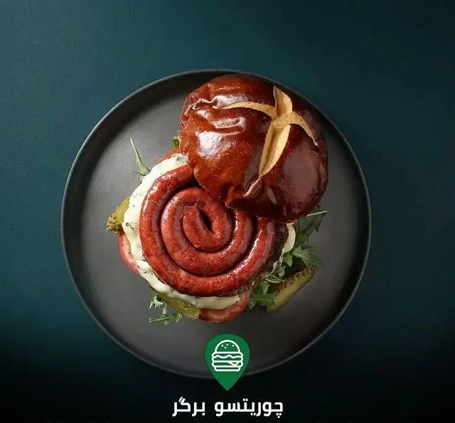 برگر چوریتسو جذاب کافه رستوران لوکال ولنجک، با سوسیس چوریتسو مارپیچ، نان پرتل و مخلفات تازه روی بشقاب مشکی.