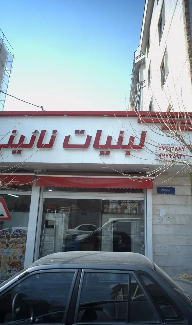 نمای خارجی فروشگاه لبنیات نائینی در تهران، با تابلوی قرمز و سفید روشن و خوانا. شامل شماره تماس ۷۷۲۹۲۸۸۷، ۷۷۲۹۴۸۷۷ و آدرس خیابان سوم نیروی هوایی.