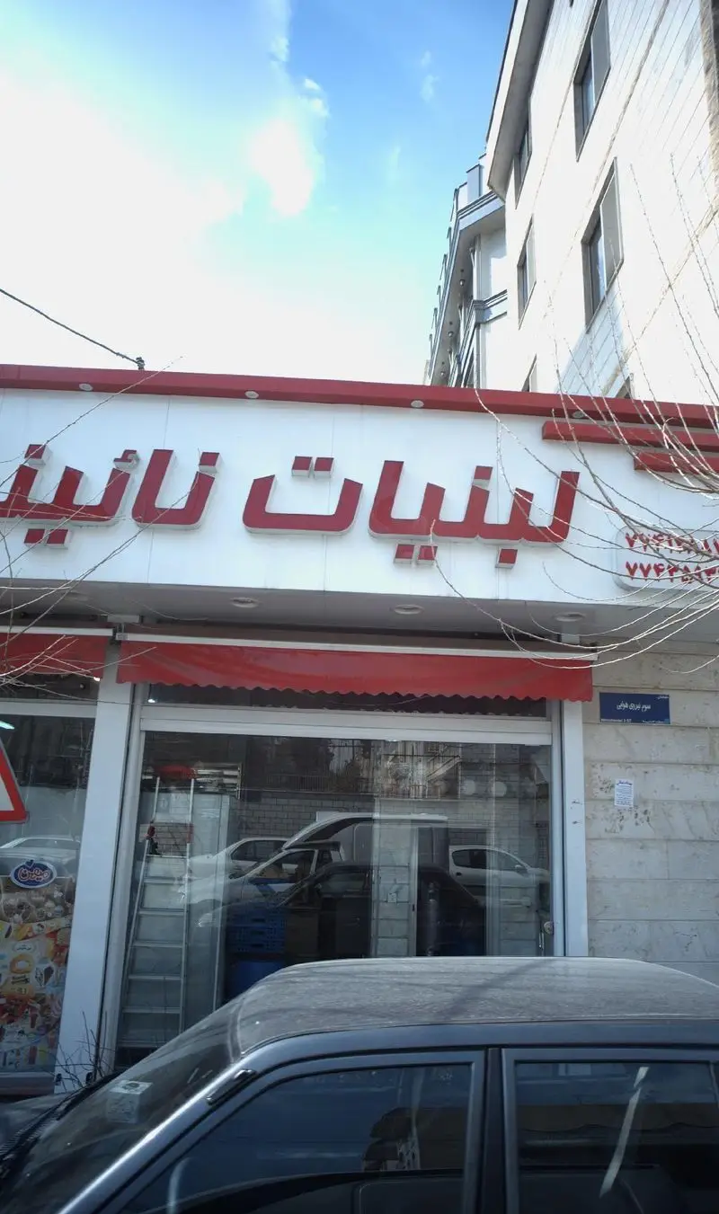 نمای بیرونی فروشگاه لبنیات نائینی با تابلوی برجسته قرمز و سفید، سایبان قرمز و ورودی شیشه‌ای شفاف در روزی آفتابی. بهترین لبنیات سنتی.