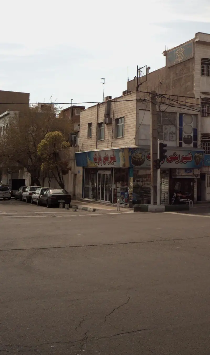 نمای بیرونی شیرینی فروشی "شیرینی پارک" در تقاطع یک خیابان شهری، با تابلوهای آبی و قرمز، خودروهای پارک شده و نمای ساختمان‌ها.