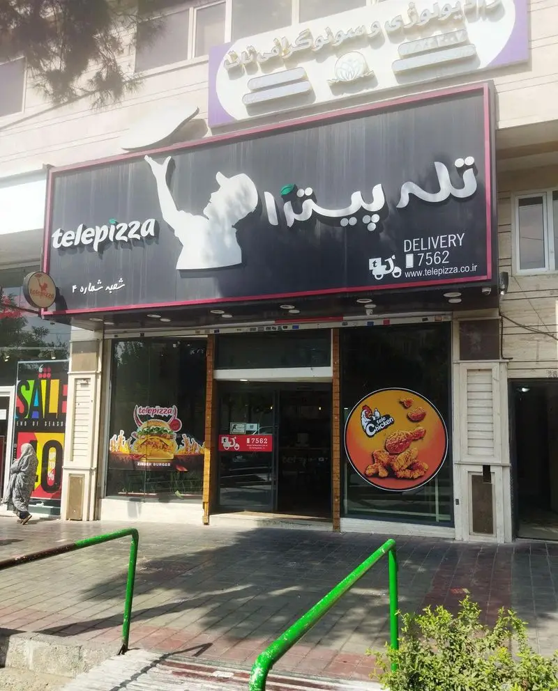 نمای بیرونی تله پیتزا شعبهی تهرانپارس با تابلوی بزرگ، لوگوی آشپز در حال پرتاب پیتزا، اطلاعات دلیوری و منوی زینگر برگر.