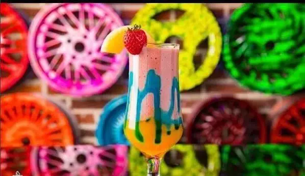 نوشیدنی لایه‌ای میوه‌ای با تزیین توت فرنگی در محیط شاد کافی شاپ پارکینگ با دکوراسیون چرخ‌های رنگی روی دیوار آجری.