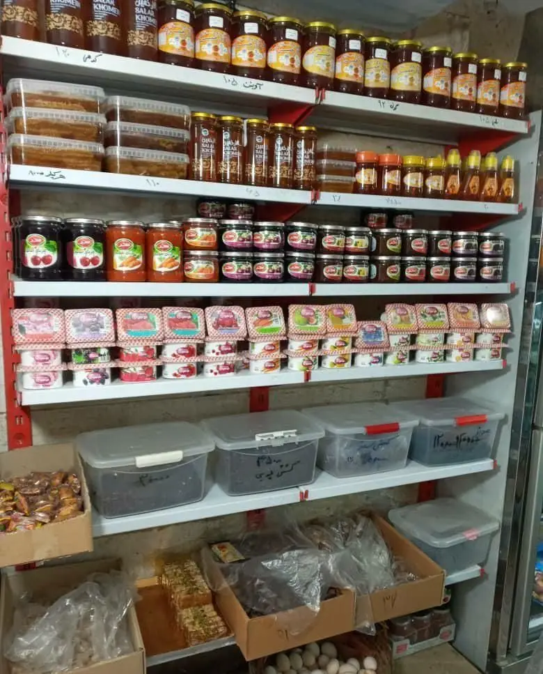 تصویری از قفسه‌های فروشگاه لبنیات سنتی دهکده با انواع عسل طبیعی، مرباهای خانگی، و محصولات محلی تازه.