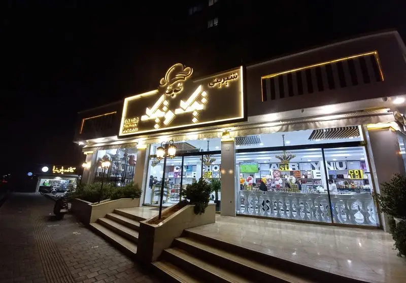 نمای بیرونی قنادی تیک و تیک در شب، با تابلوی روشن و ویترین پر از انواع شیرینی و دسر، آماده پذیرایی از مشتریان.