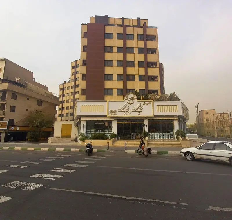 نمایی بیرونی از قنادی تیک و تیک با ویترین‌های جذاب، تابلوی بزرگ و ساختمان آپارتمانی بلند در پس‌زمینه، در یک روز ابری.