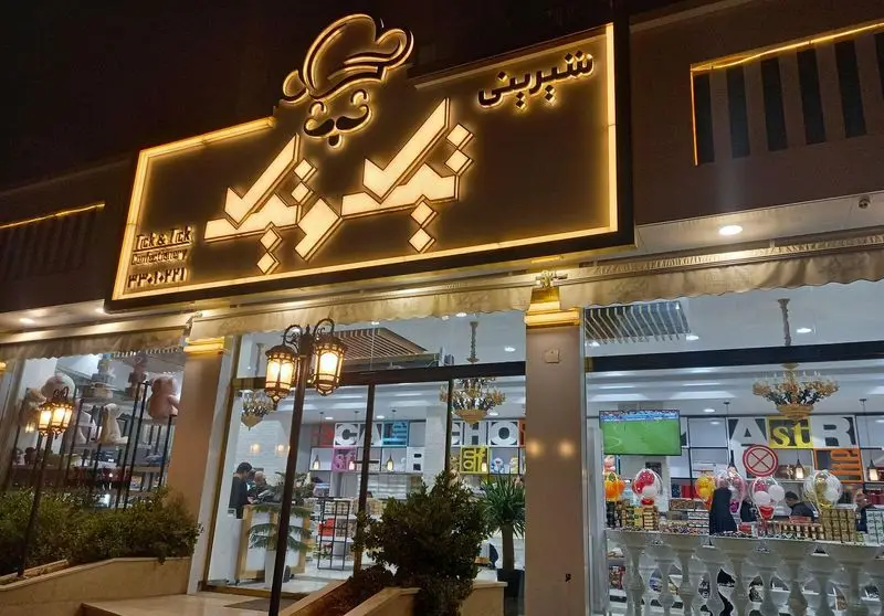 تصویری جذاب از نمای شبانه قنادی تیک و تیک، شیرینی فروشی لوکس با نورپردازی زیبا و انواع کیک، دسر و شکلات.